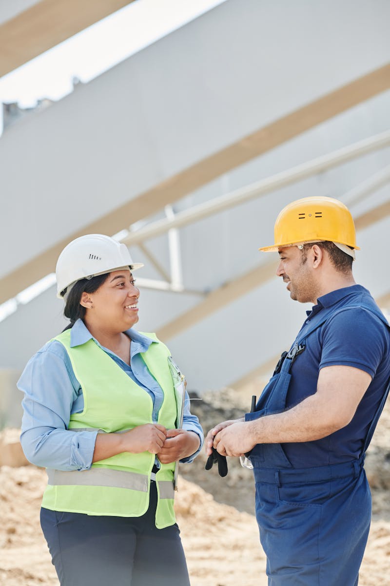 Two construction workers wearing safety gear discussing plans outdoors at a construction site.